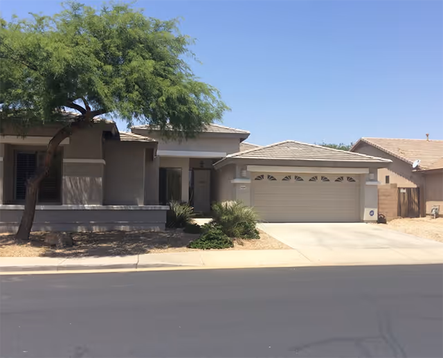 Single-story stucco house with a two-car garage, driveway, and desert landscaping under a clear blue sky.