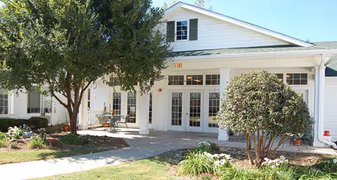 Front entrance of a white senior living building with double glass doors, columns, porch seating, and landscaped trees and shrubs.