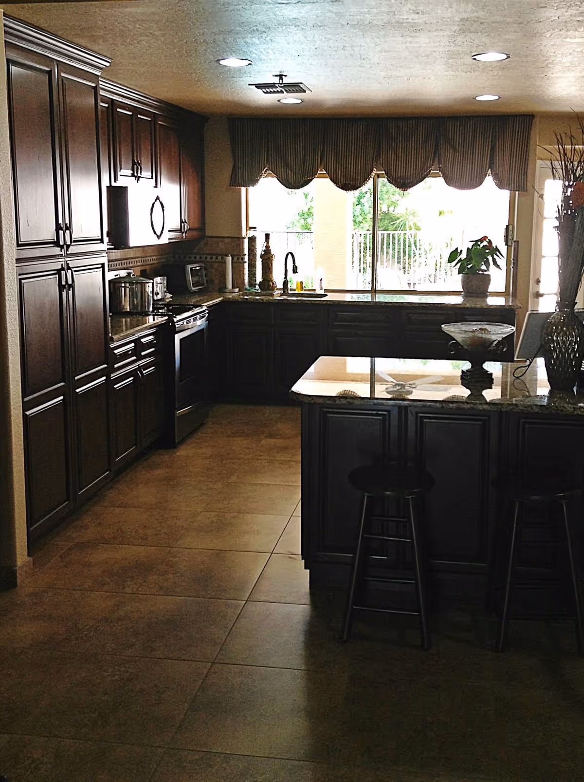 A kitchen with dark wooden cabinets, a stove, a microwave, and a kitchen island with two stools. There is a large window with a valance above the sink, and various decorative items on the countertops.