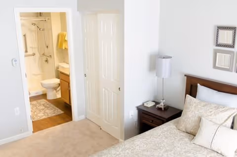 View of a bedroom with a bed, pillows, and a nightstand with a lamp. The room has light-colored walls and carpet. An open door reveals a bathroom with a toilet, shower, and yellow towels.