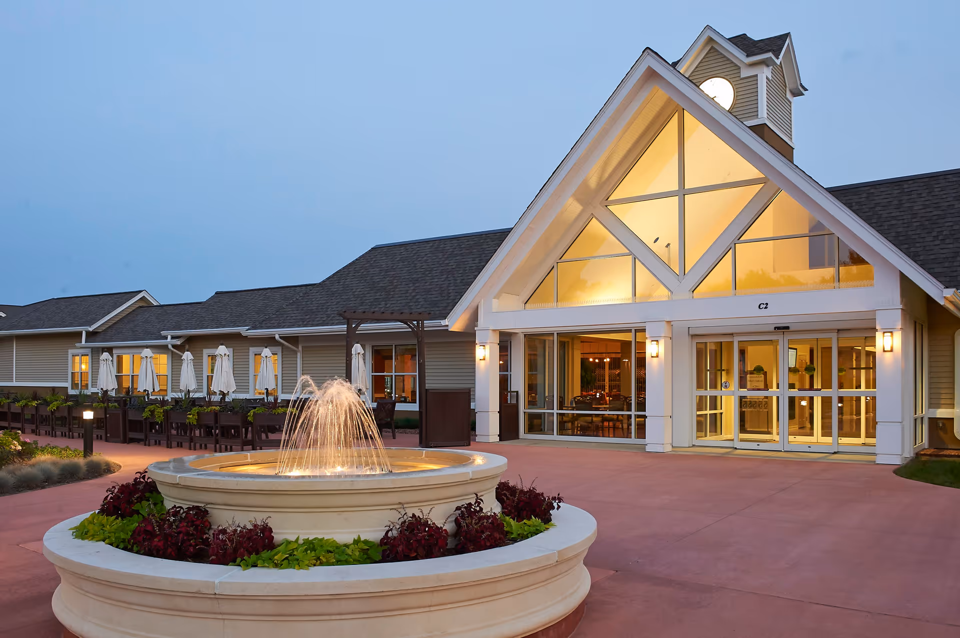 Exterior view of a senior living facility building at dusk with a large, illuminated fountain in the foreground. The building has a peaked roof with large windows and warm interior lighting visible through the glass doors and windows. There are outdoor seating areas with tables and umbrellas along the side of the building.