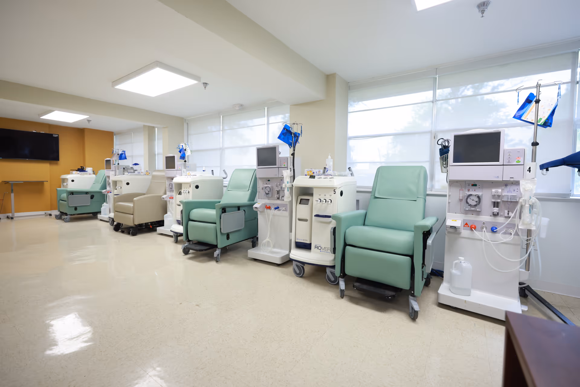 A clean and well-lit medical treatment room with several reclining chairs and dialysis machines lined up against a wall with large windows covered by white blinds. The floor is shiny and beige, and there is a large flat-screen TV mounted on a mustard-colored wall in the background.