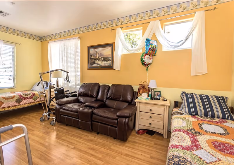 A cozy room with two single beds on either side, each covered with colorful patchwork quilts. Between the beds is a brown leather reclining sofa and a small wooden nightstand with a lamp, tissue box, and framed photo. The walls are painted yellow with a floral border near the ceiling. Two small windows with sheer white curtains let in natural light. A walker and a patient lift device are also visible in the room.