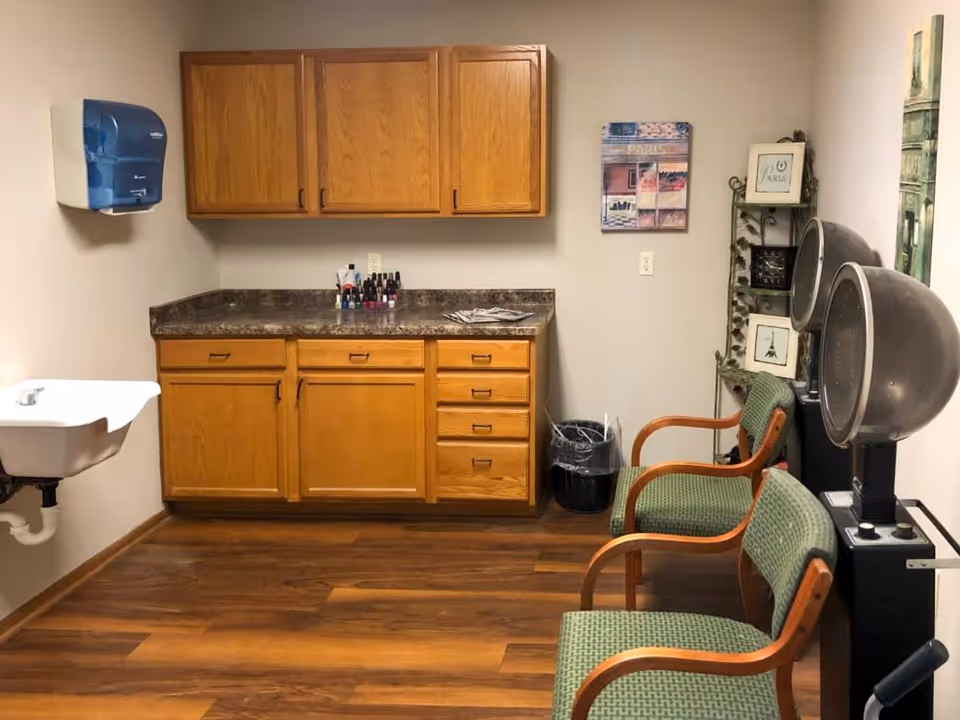 Interior room with wooden cabinets and countertop, a small white sink on the left wall, two green cushioned chairs with wooden armrests, and two vintage hair dryers. The room has wood flooring, a wall-mounted paper towel dispenser, a trash can, and framed Paris-themed wall art.