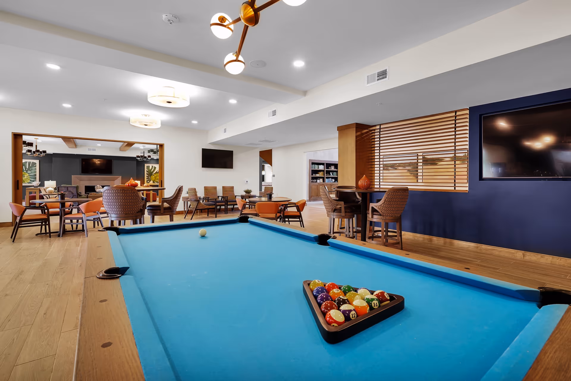 A bright communal game room with a blue-felt pool table in the foreground and seating areas, tables, and TVs in the background.