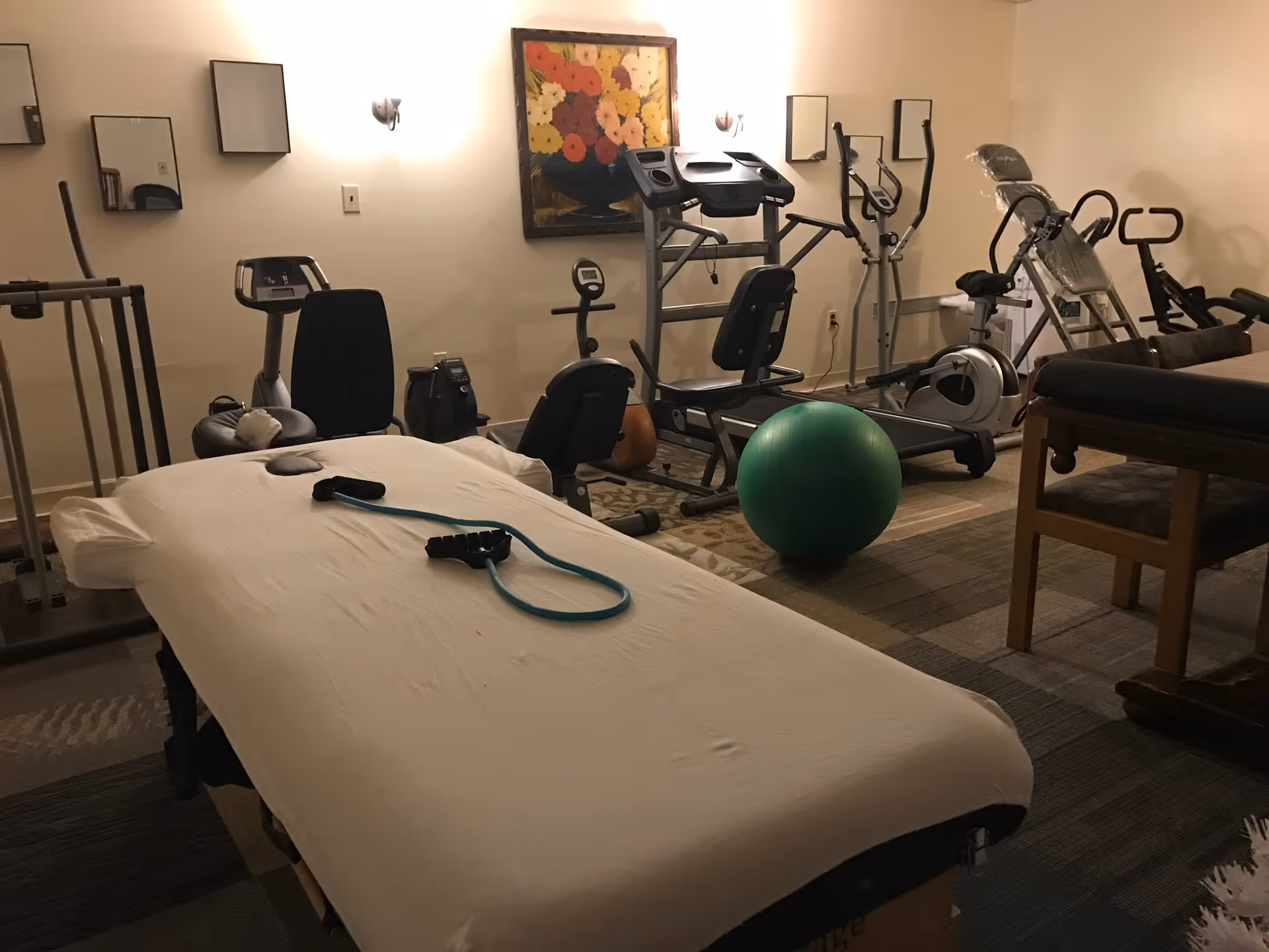 A room with various exercise equipment including a treadmill, stationary bikes, elliptical machines, and a green exercise ball. In the foreground, there is a massage table with a white sheet and a resistance band on top. The walls are beige with a floral painting and two small mirrors. The room has a carpeted floor and soft lighting.
