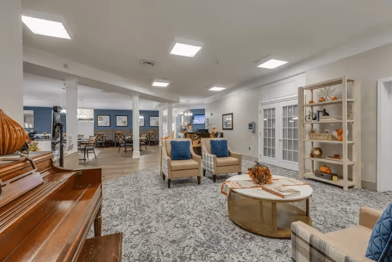 A spacious and well-lit common area in a senior living facility featuring a wooden piano on the left, two beige armchairs with blue cushions, a round coffee table decorated with autumn-themed items, and a white shelving unit with seasonal decorations. In the background, there is a dining area with tables and chairs, white columns, and a television mounted on the wall.