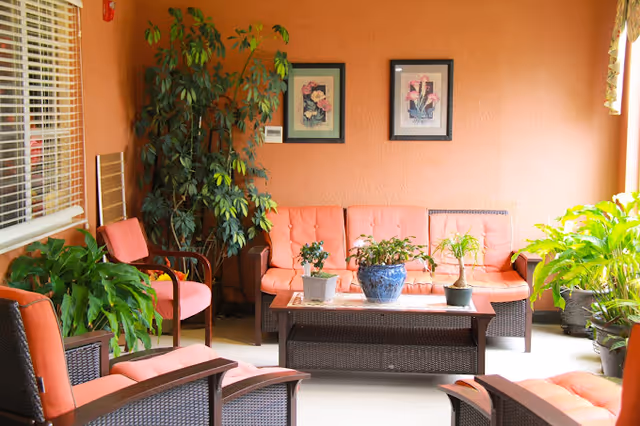 Bright seating area with coral-upholstered wicker chairs and sofa around a coffee table with potted plants, framed artwork on peach walls and many green plants.
