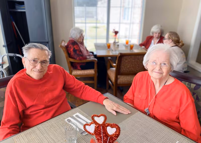 An elderly man and woman sitting at a dining table in a senior living facility, both wearing red tops. The man is smiling and wearing glasses, while the woman is also smiling and wearing a necklace. In the background, three other elderly women are seated at another table near a large window with drinks on the table.