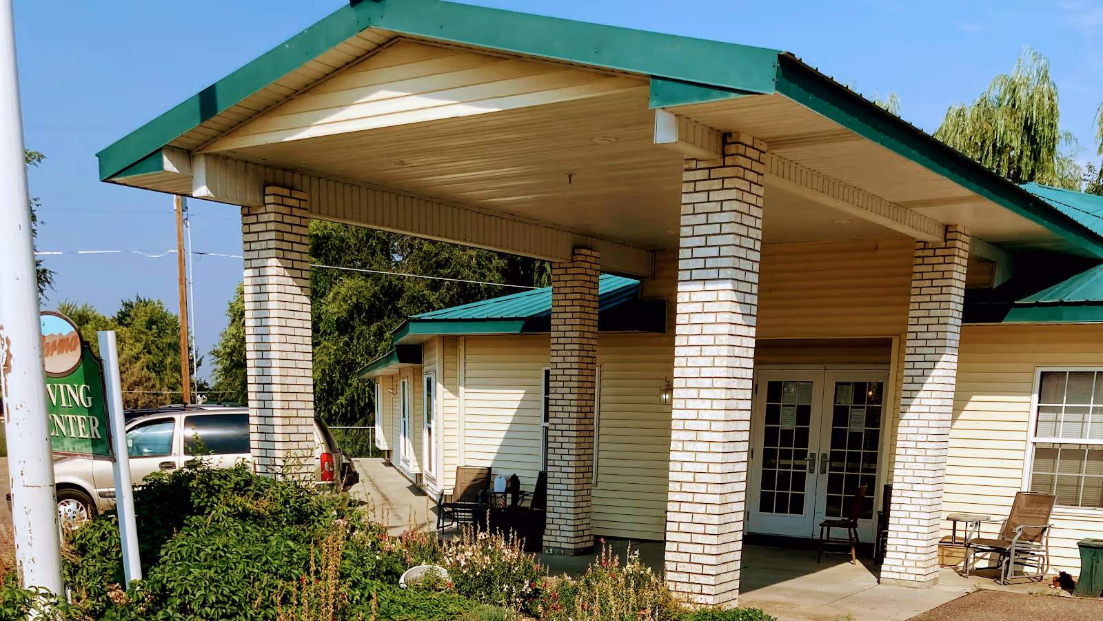 Covered front entrance of a single-story senior living building with brick pillars, a green roof, and a parking area to the left.