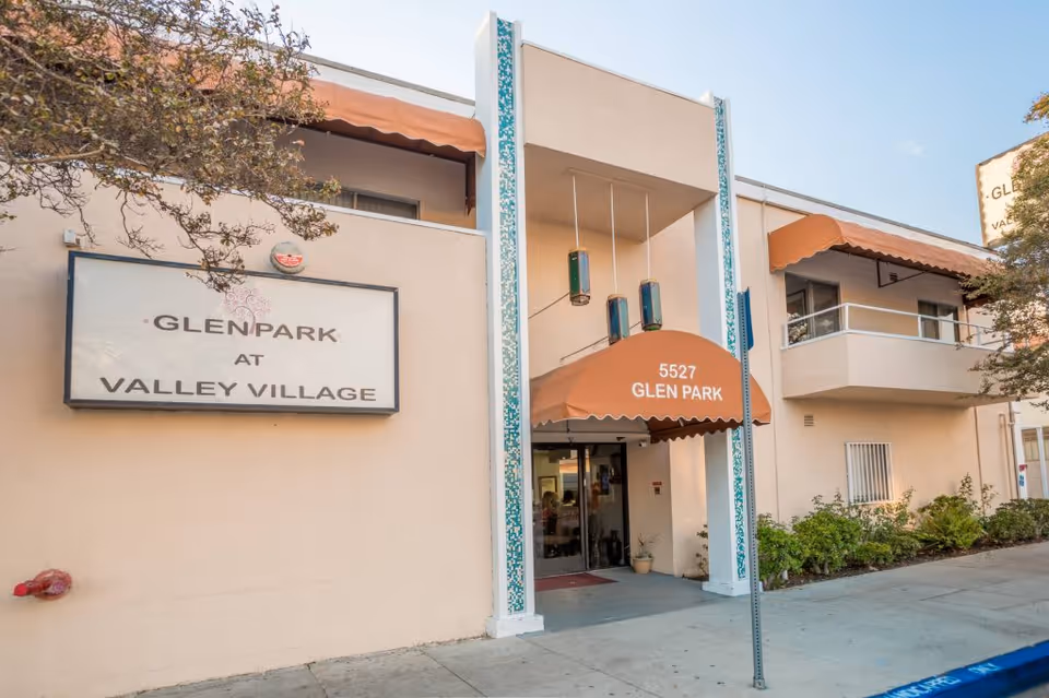 Exterior view of Glen Park at Valley Village senior living facility showing the entrance with an orange awning labeled '5527 Glen Park' and a large sign on the building that reads 'Glen Park at Valley Village'. The building is beige with balconies and some greenery around the entrance.