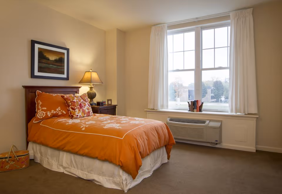 A cozy bedroom with a single bed covered in an orange bedspread and decorative pillows. There is a wooden headboard, a nightstand with a lamp, and a framed landscape painting on the wall. A large window with white curtains lets in natural light, and an air conditioning unit is installed below the window.