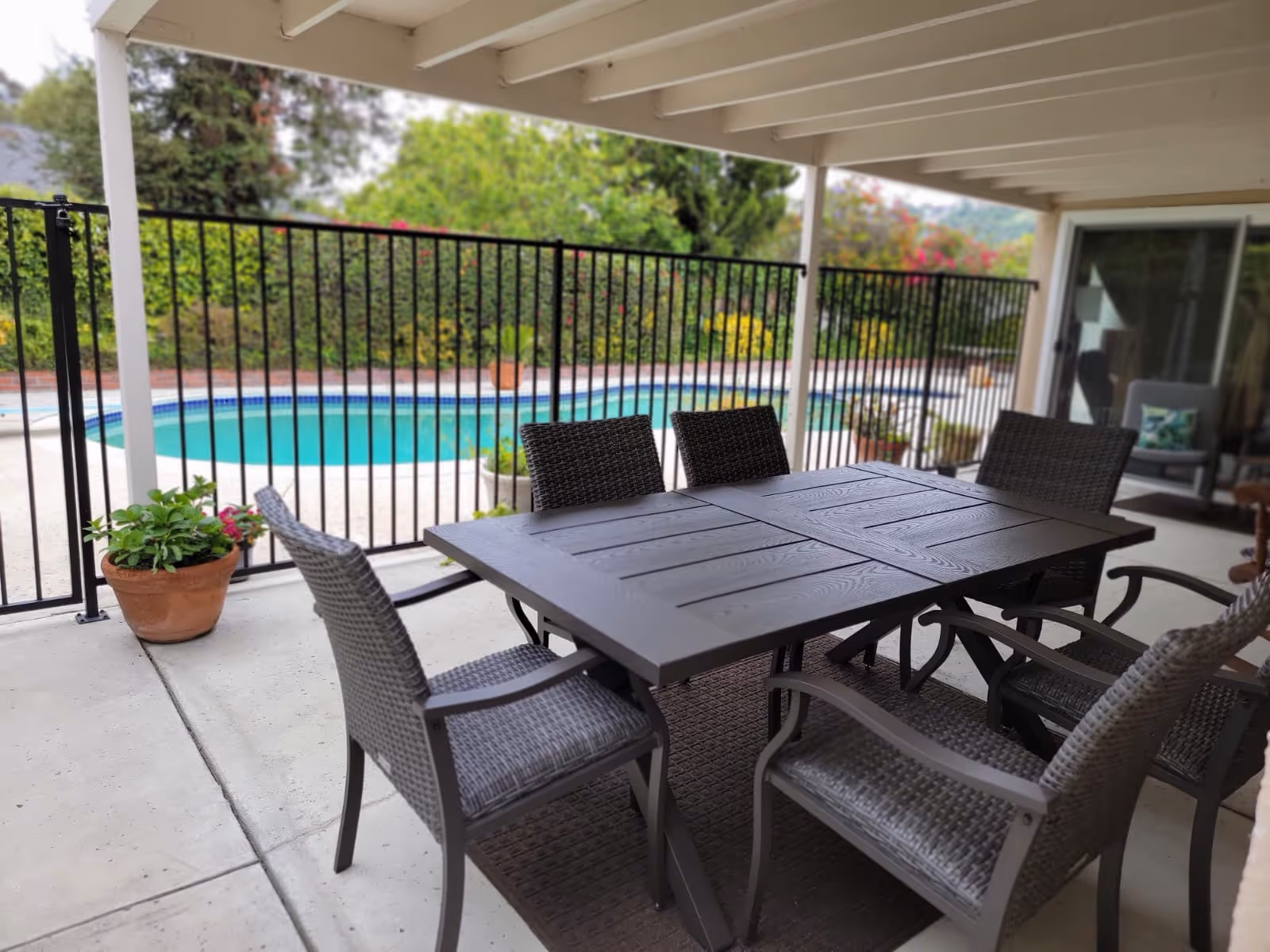 Outdoor covered patio area with a dark rectangular table and six wicker chairs. Beyond the black metal fence is a swimming pool surrounded by greenery and plants.