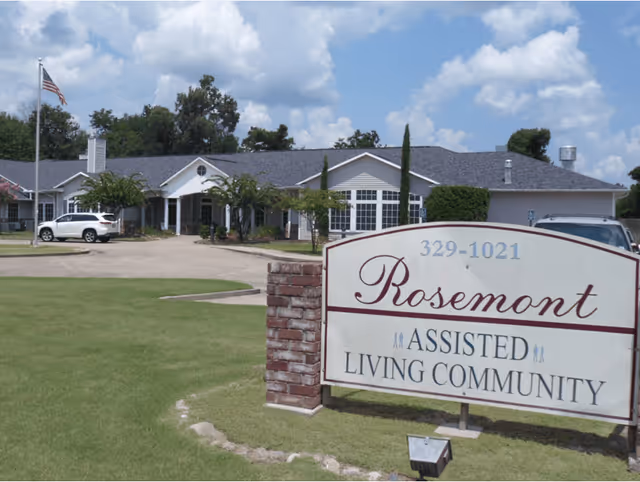 Front view of the Rosemont assisted living community sign with the single-story facility, driveway, lawn, and parked cars in the background.