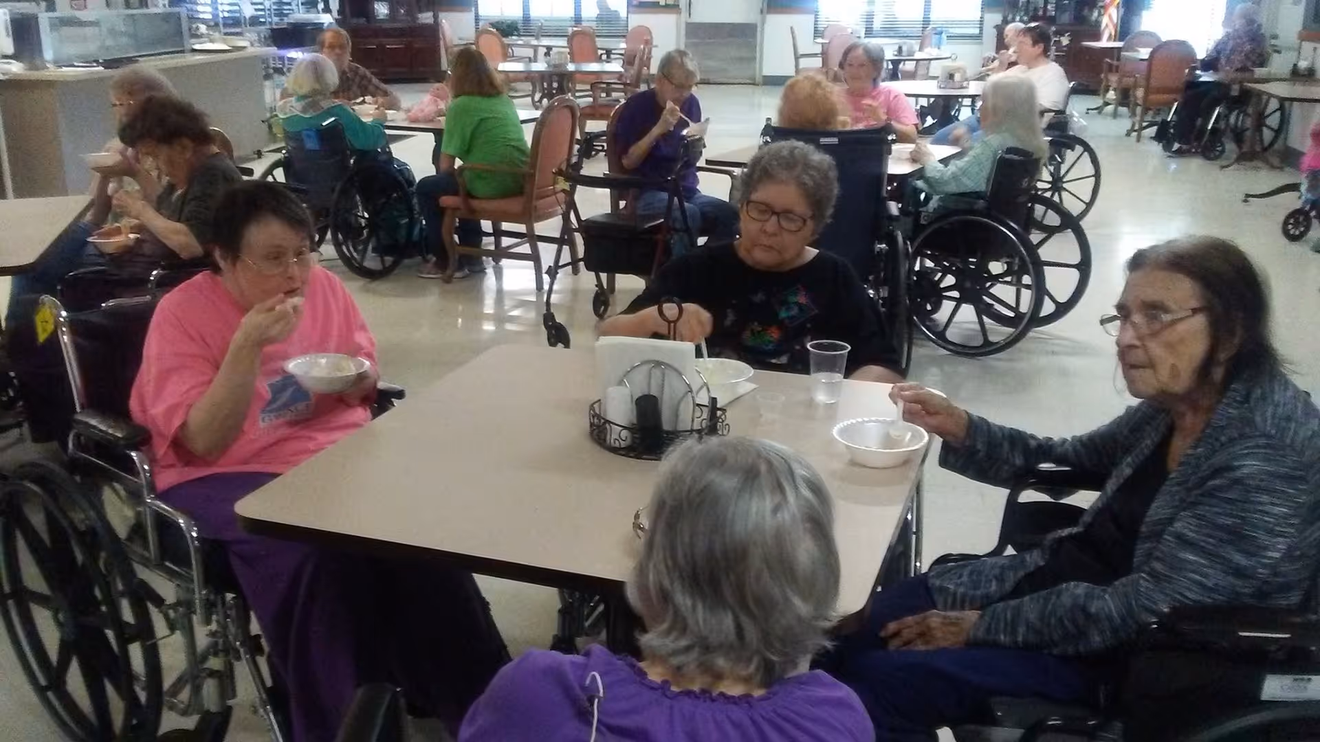 Several elderly residents in wheelchairs sit around tables in a communal dining area eating from bowls.