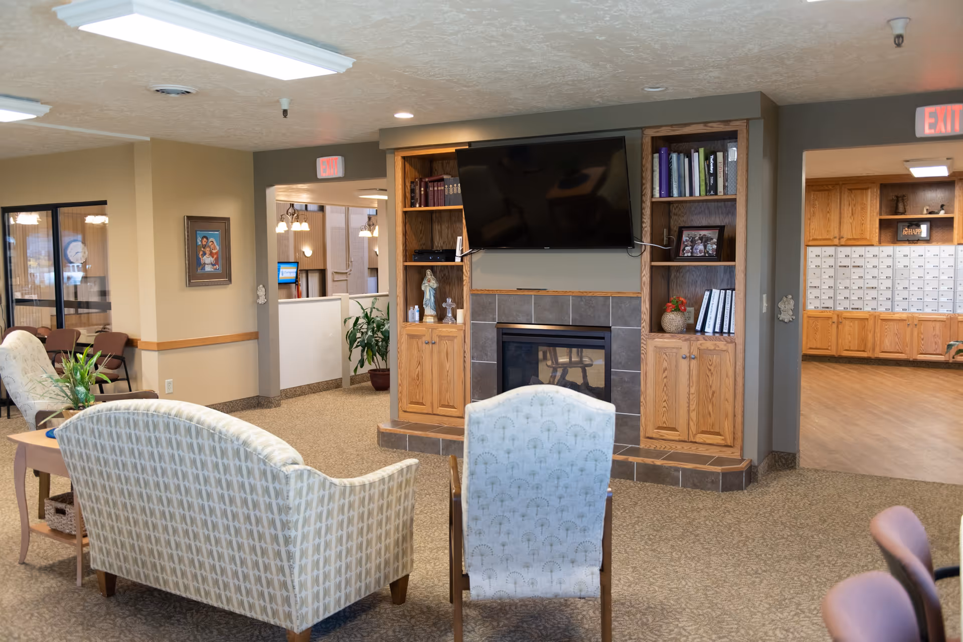 A cozy senior living facility common area with patterned upholstered chairs and a loveseat facing a wall-mounted flat screen TV above a fireplace. Wooden built-in shelves flank the fireplace, holding books, decorations, and framed photos. The room has beige walls, carpeted floors, and overhead fluorescent lighting. Adjacent rooms are visible, including one with mailboxes and another with a seating area and a framed picture on the wall.