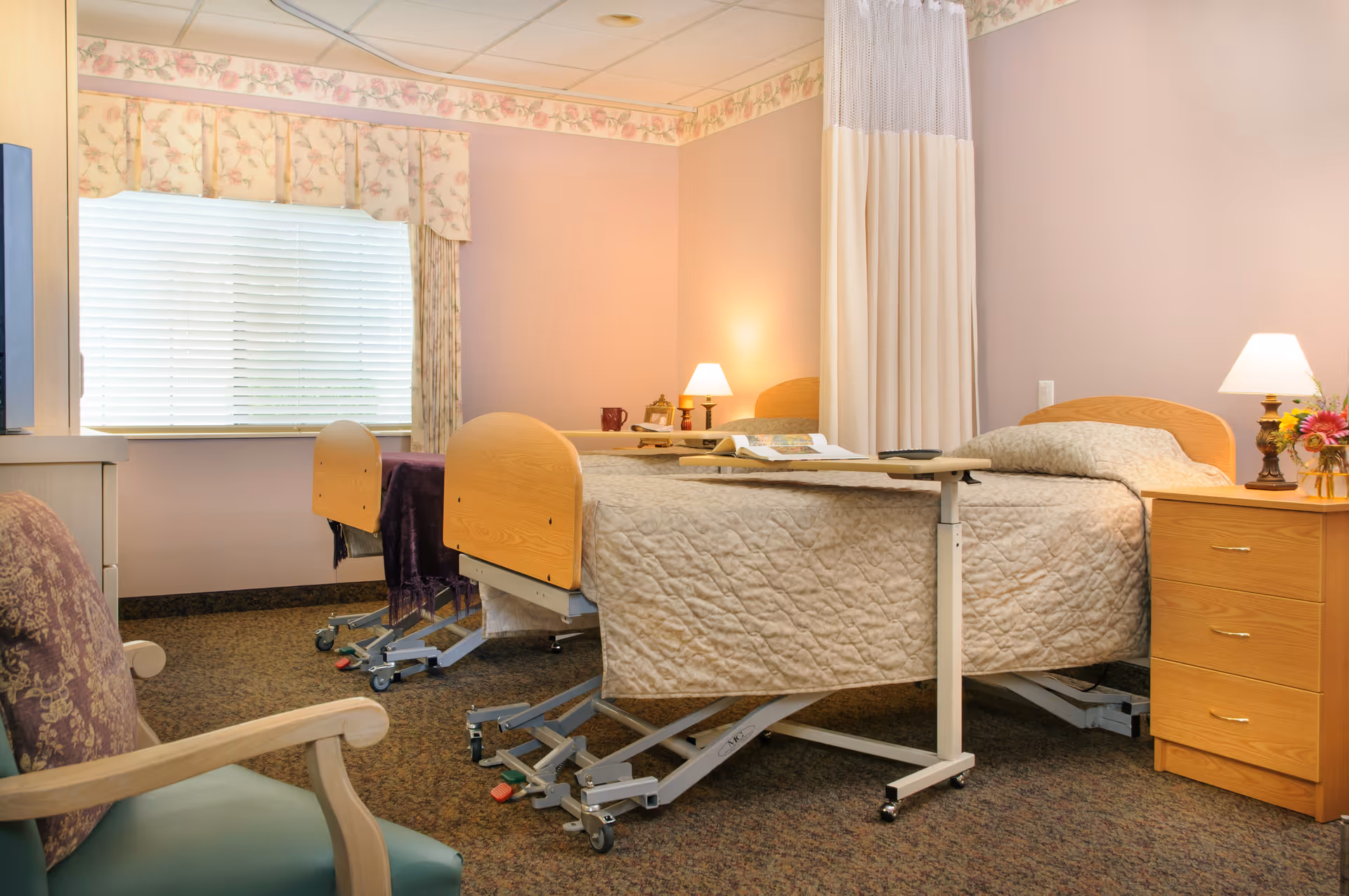 Two adjustable hospital-style beds in a softly decorated senior living bedroom with bedside tables, lamps, and a window.