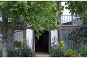 Entrance to a residential building with a pathway leading through a garden area with various plants and trees. The building has large windows and a balcony above the entrance.