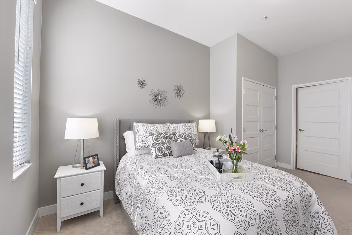 Bright bedroom with a neatly made bed featuring patterned bedding, bedside tables with lamps, wall decor, and a tray of flowers.