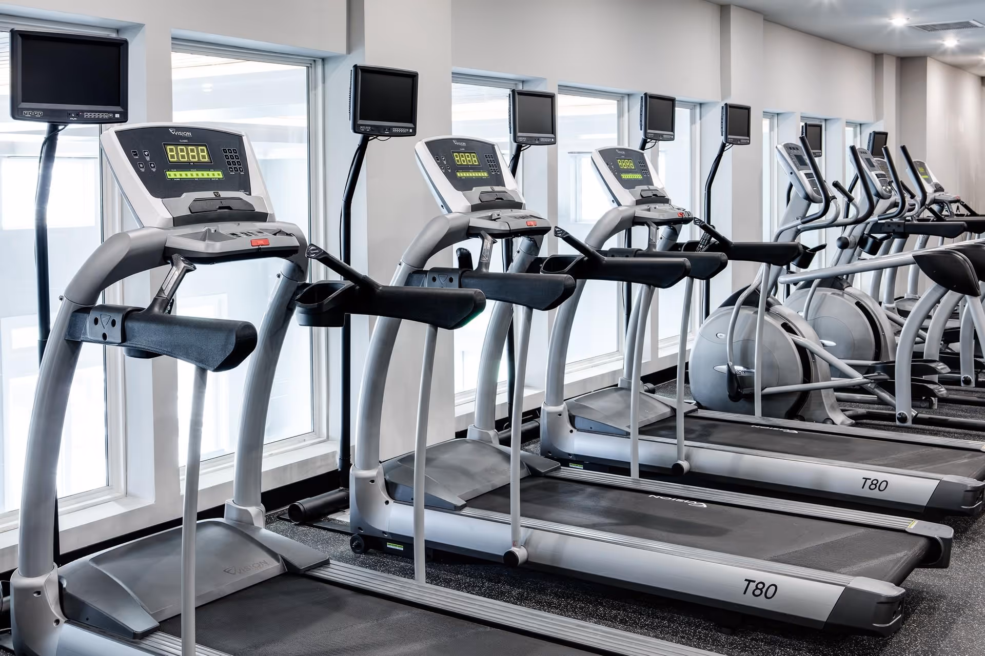 A row of modern treadmills and elliptical machines in a bright fitness room with large windows allowing natural light to enter.