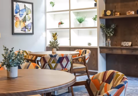 A cozy interior room with round wooden tables and colorful geometric-patterned chairs. The background features a white shelving unit with potted plants and a framed butterfly artwork on the wall. There is also a wooden desk area with decorative items and a digital clock.