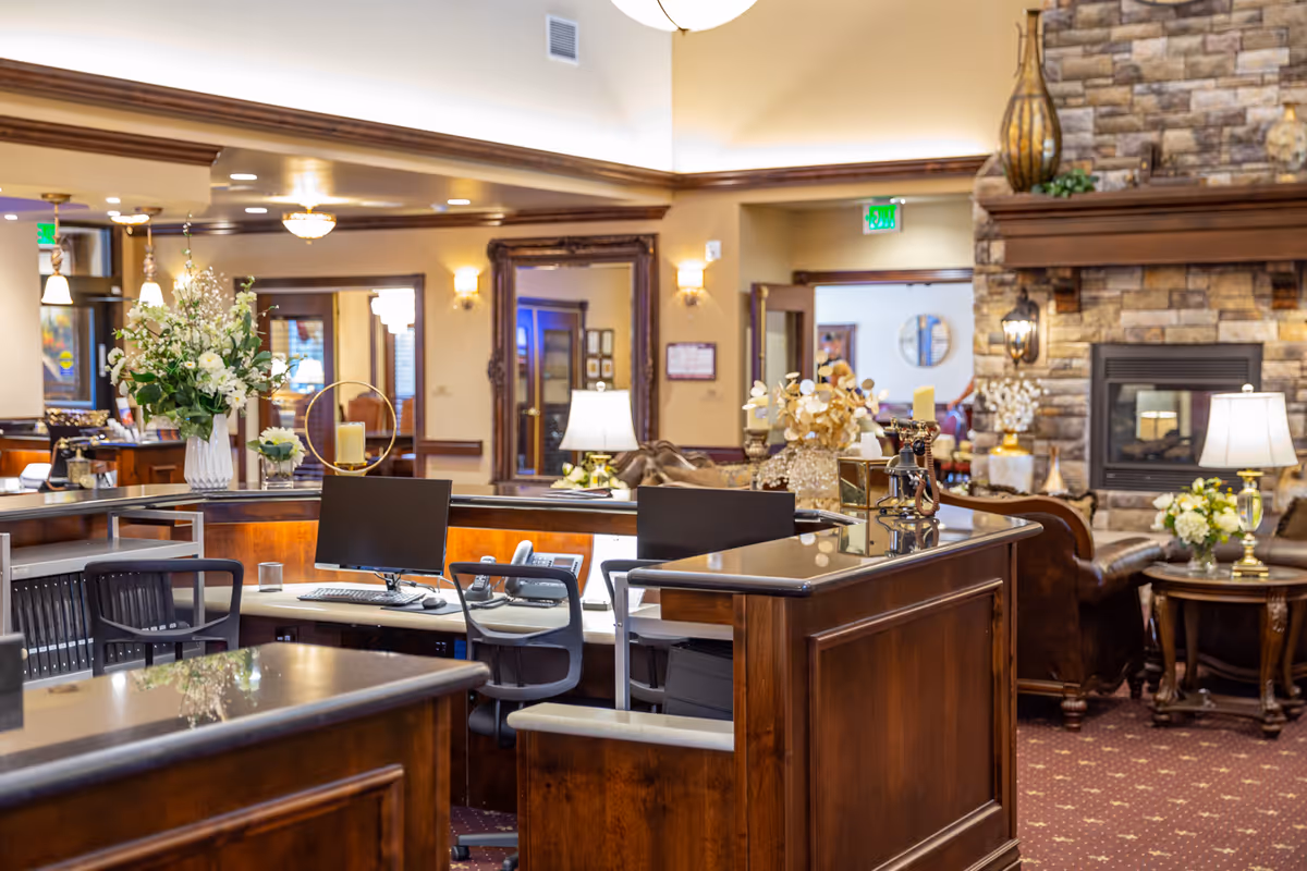 Reception area with a wooden desk featuring computer monitors and office chairs. Behind the desk is a cozy lounge area with leather chairs, a stone fireplace, and decorative flowers and lamps. The space is warmly lit with ceiling lights and wall sconces.