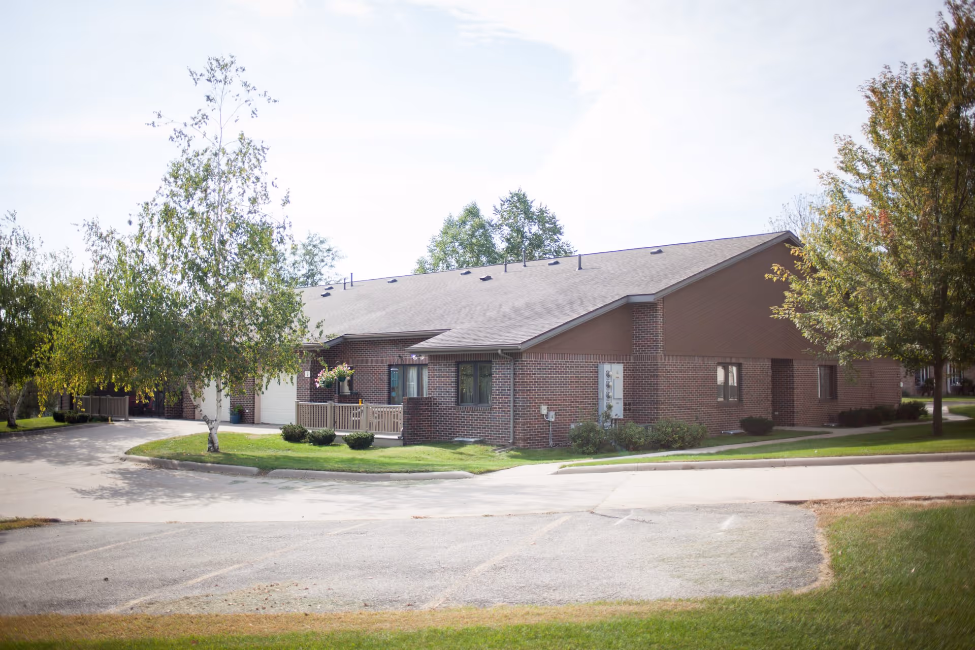 Exterior view of a single-story brick building with a sloped roof, surrounded by trees and a paved driveway, under a partly cloudy sky.