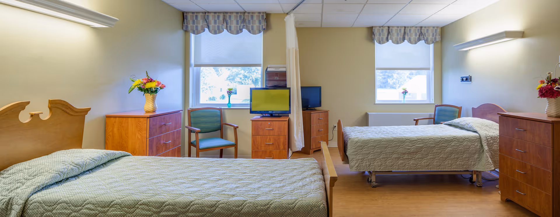 A bright and clean shared bedroom in a senior living facility with two single beds, each with a green quilted bedspread. Between the beds are wooden dressers topped with flower vases. Two chairs are positioned near windows with patterned valances, and two small televisions sit on dressers. The room has light-colored walls and wood flooring.