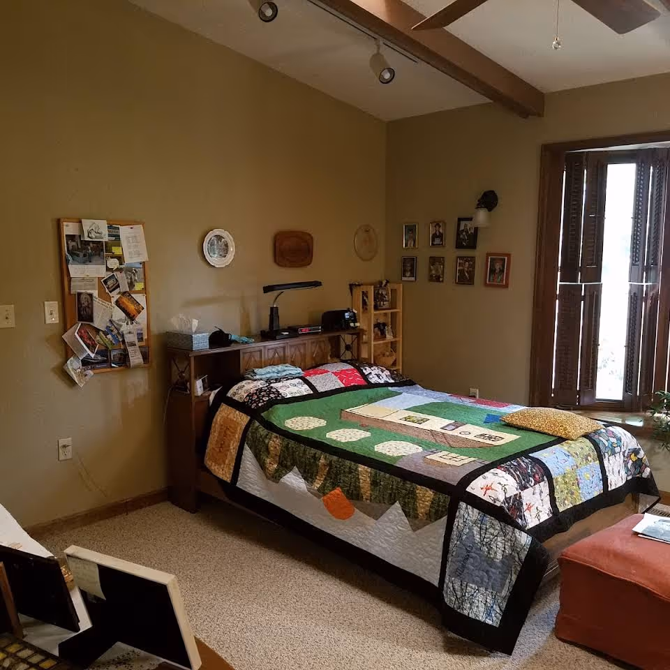 A cozy bedroom with a colorful patchwork quilt on the bed, a bulletin board and framed photos on the walls, and a window with wooden shutters.