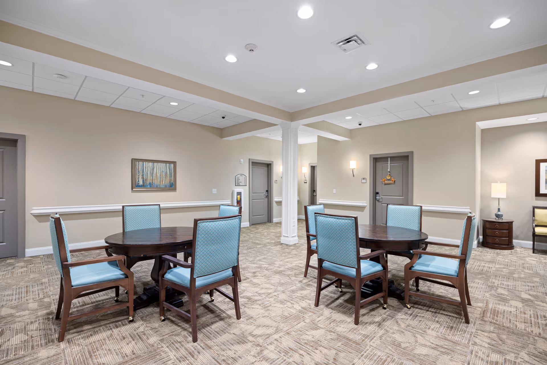 A well-lit interior room with two round wooden tables, each surrounded by four blue cushioned chairs. The room has beige walls, carpeted flooring, and several closed gray doors. There is a framed painting on one wall and a small table with a lamp and a chair in a recessed area. The ceiling has recessed lighting and a white support column in the center.