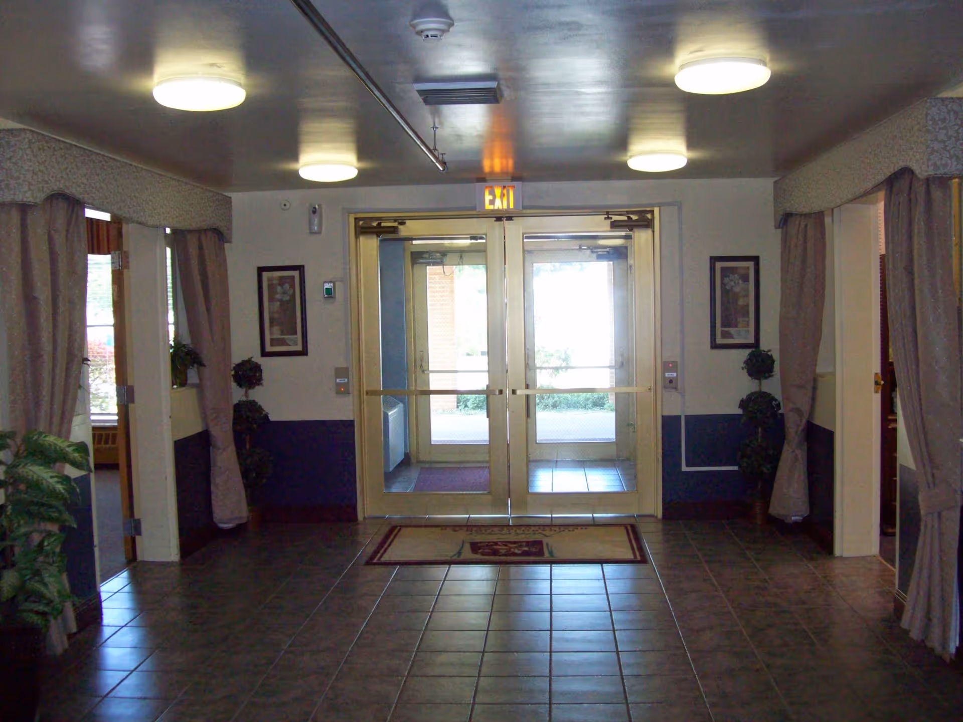 Interior view of an entrance lobby with double glass doors and an exit sign above. The floor is tiled, and there are framed pictures and potted plants on either side of the doors. Curtains hang on the walls beside doorways leading to other rooms.
