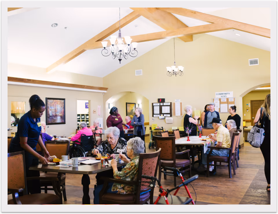 A bright and spacious common dining area in a senior living facility with wooden beams on the ceiling and chandeliers. Several elderly residents are seated at tables eating and socializing, while staff members assist them. The room has warm yellow walls, framed artwork, and a community news board on the far wall.