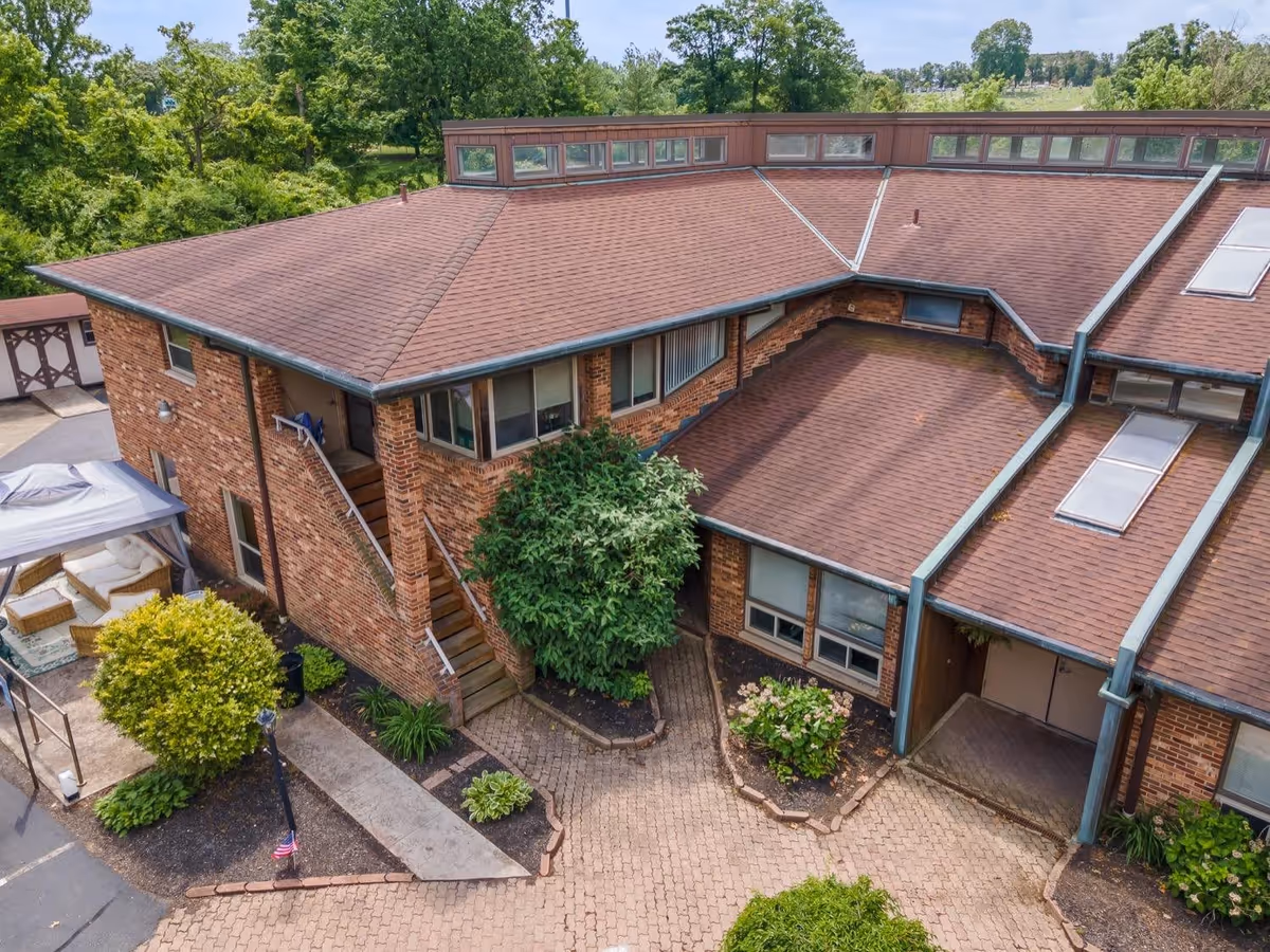 Aerial view of a brick building with a brown shingled roof surrounded by greenery. The building has multiple windows, an outdoor staircase, and a small paved courtyard with plants and shrubs. There is also a covered outdoor seating area with wicker furniture and cushions visible on the left side.