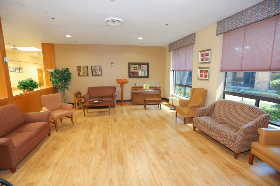 A spacious and well-lit living room area with wooden flooring, multiple upholstered chairs and sofas arranged around the room, large windows with blinds letting in natural light, a piano against the far wall with a small bench, framed artwork on the walls, and a potted plant in the corner.