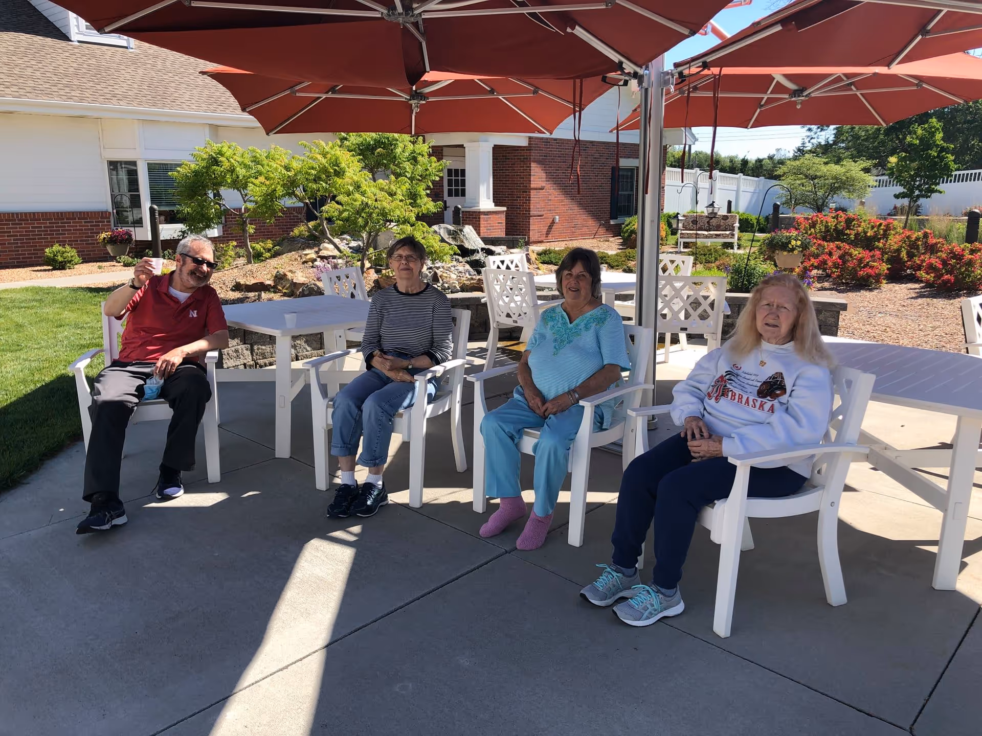Four elderly people sitting on white chairs under large red umbrellas in an outdoor patio area with tables, greenery, and a brick building in the background.