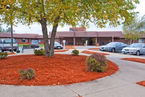 Front entrance of a single-story rehabilitation and health care center with a circular driveway, landscaped mulch beds and parked cars.