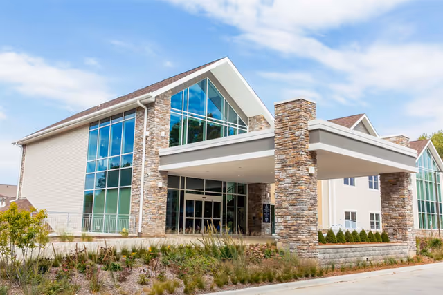 Exterior view of a modern senior living facility building with large glass windows, stone pillars, and a covered entrance. The building is surrounded by landscaped greenery and a clear blue sky is visible above.