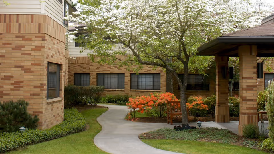 Outdoor garden area at West Hills Village Senior Residence featuring a curved concrete walkway, a tree with white blossoms, orange flowering bushes, green shrubs, and a covered seating area with wooden benches supported by brick pillars.