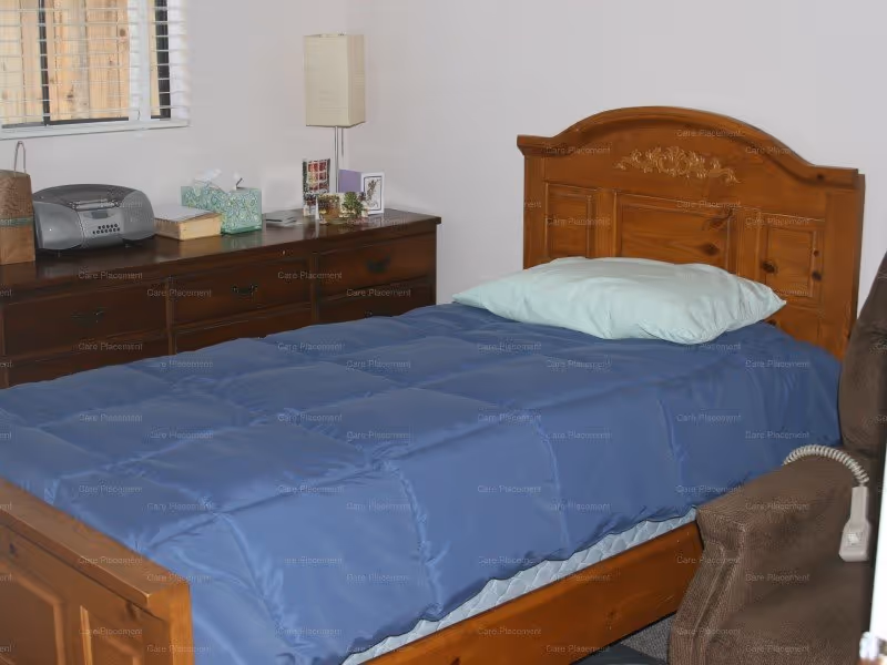 A bedroom with a wooden bed frame and a blue comforter, a light blue pillow, a wooden dresser with various items on top including a radio, tissue box, and lamp, and a brown upholstered chair with a white corded remote control.