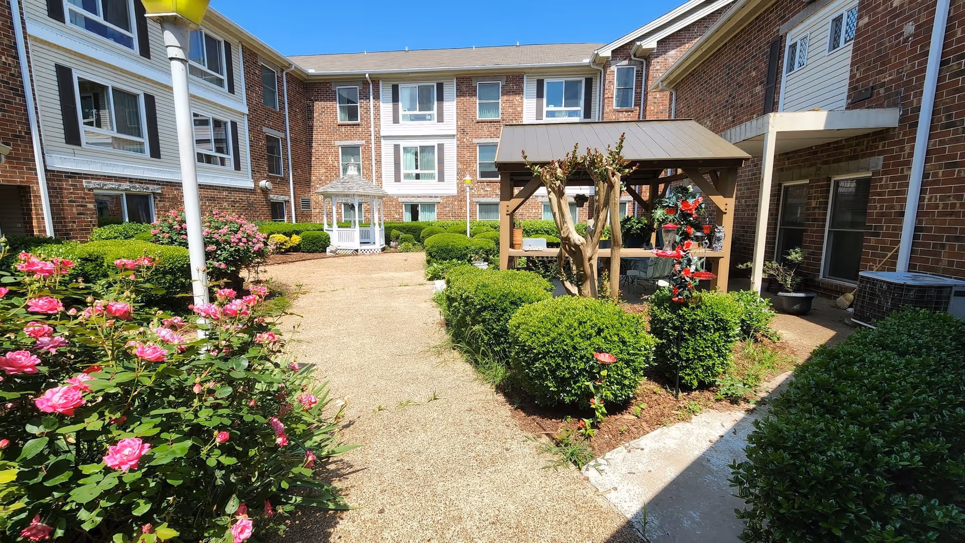 Outdoor courtyard area of a senior living facility with a pathway surrounded by green bushes and blooming pink roses. There is a white gazebo in the background and a wooden pergola with seating on the right side. The building is made of red brick with multiple windows, and the sky is clear and blue.