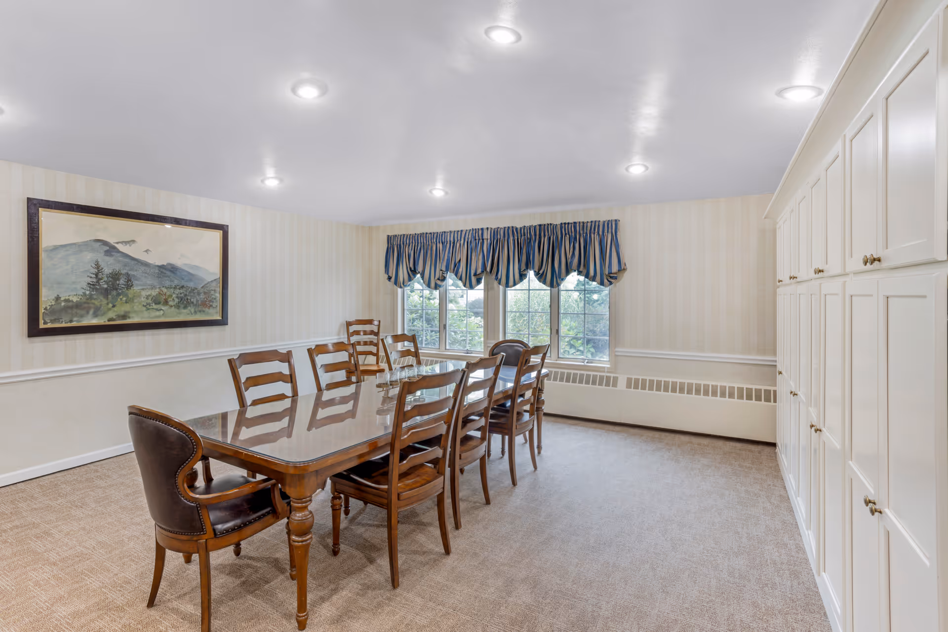 A dining room with a long wooden table surrounded by eight wooden chairs, one of which has a leather backrest. The room has beige carpet flooring, cream-colored wallpaper with subtle stripes, a large window with blue and beige striped valances, and a large white cabinet along one wall. A framed landscape painting hangs on the opposite wall.