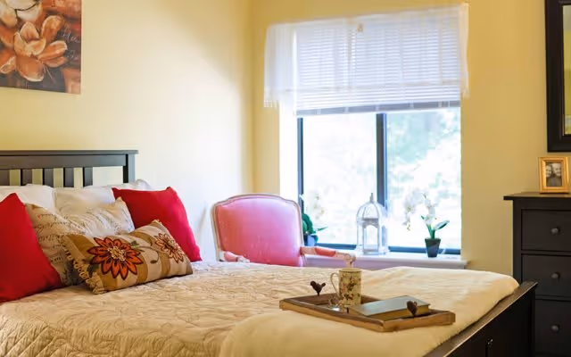 A cozy bedroom with a neatly made bed featuring a beige quilt, red and patterned pillows, and a wooden tray with a mug and book on top. There is a pink upholstered chair near a window with white blinds, and a dark wooden dresser with a framed photo on it. A floral painting hangs on the wall above the bed.