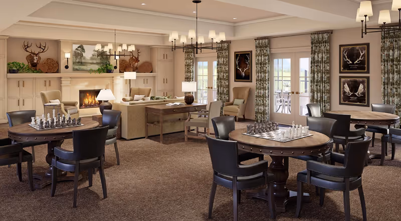 A cozy senior living community room with multiple round tables set up for chess games, surrounded by black leather chairs. The room features a lit fireplace with decorative items and paintings above it, beige armchairs, a sofa, and large windows with floral curtains letting in natural light.