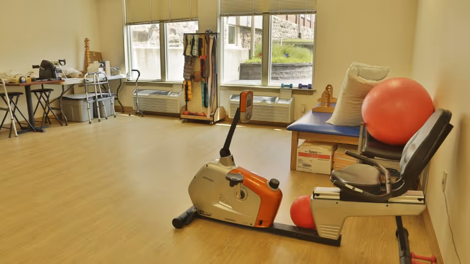 A physical therapy or exercise room with a recumbent exercise bike, a large red exercise ball, a smaller red ball, a padded bench with pillows, and various therapy equipment including a walker, weights, and resistance bands near windows with blinds.