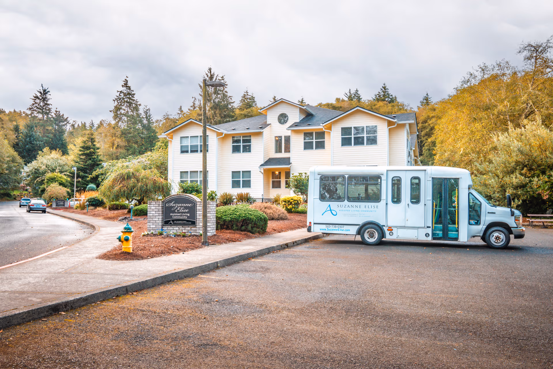 The Suzanne Elise Assisted Living Community building, a white two-story facility, with a light-blue bus parked in front.