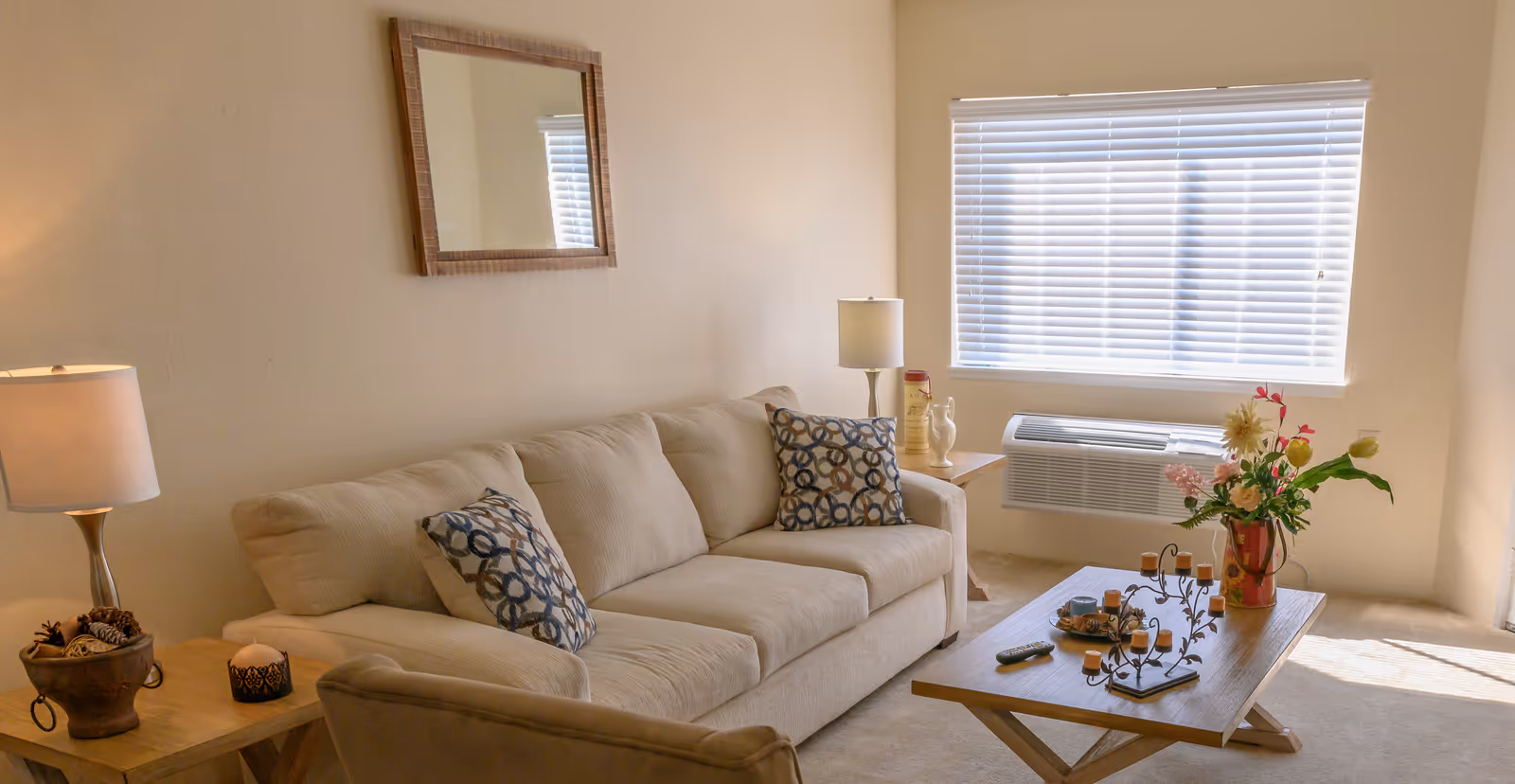 A cozy living room with a beige sofa adorned with two patterned cushions, a wooden coffee table with decorative items and a remote control, a side table with a lamp and small decor, a window with blinds, and a wall-mounted air conditioning unit below the window.