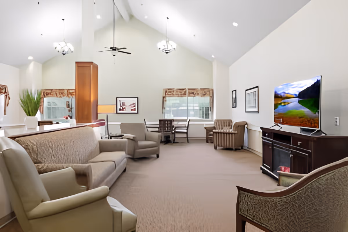 A spacious living room with high vaulted ceilings, beige carpet, and multiple seating options including sofas and armchairs. The room features a large flat-screen TV on a dark wooden stand, several framed pictures on the walls, two ceiling fans, and large windows with patterned valances allowing natural light to enter.