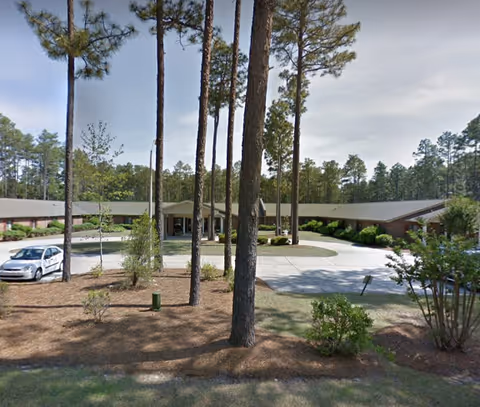 Front entrance of a single-story senior living facility with a circular driveway and tall pine trees in the landscaped front yard.