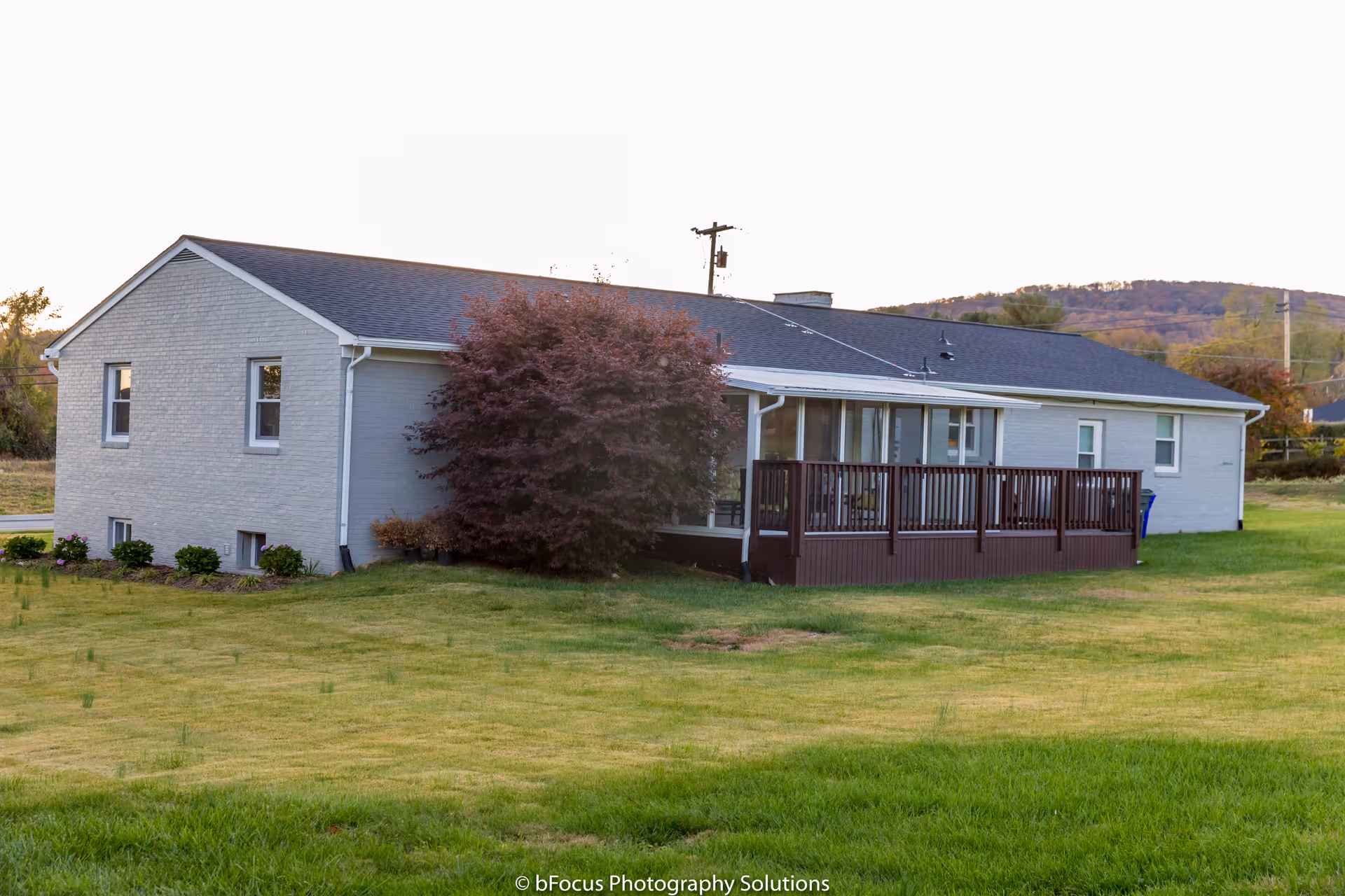 Single-story light gray brick building with a dark shingled roof, a wooden deck with railings, and a large bush near the side. The building is surrounded by a grassy lawn with hills in the background under a clear sky.
