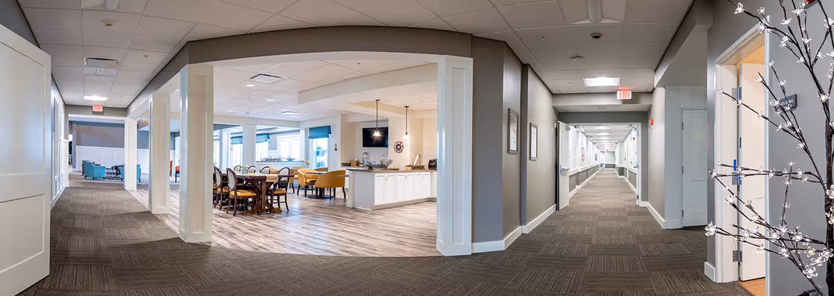Interior view of a senior living facility hallway with carpeted floors and neutral-colored walls. To the left, there is an open area with a dining table and chairs, a small kitchen or serving counter, and seating with armchairs. The hallway extends straight ahead with several doors along the walls. A decorative branch with small lights is visible on the right side near an open door.